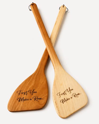 Two wooden paddle-shaped utensils crossed over each other on a light gray background, with engraved script text reading 'First You Make a Roux.