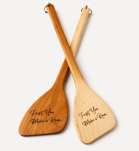 Two wooden paddle-shaped utensils crossed over each other on a light gray background, with engraved script text reading 'First You Make a Roux.