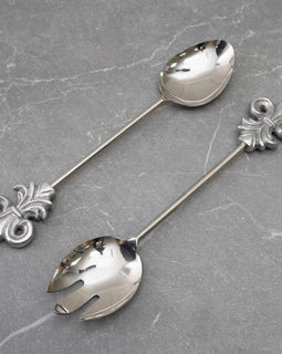 Stainless steel salad server set with fleur de lis handles on gray stone background.