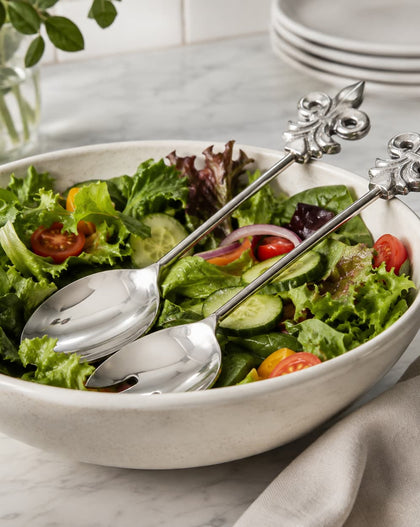 Two silver salad servers with ornate handles resting in a bowl of mixed green salad on a light marble surface.