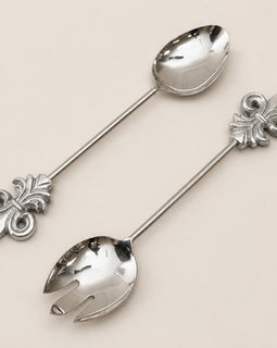 Two polished silver serving utensils with decorative fleur-de-lis handles, one spoon and one fork, arranged diagonally on a light beige background.