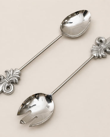 Two polished silver serving utensils with decorative fleur-de-lis handles, one spoon and one fork, arranged diagonally on a light beige background.