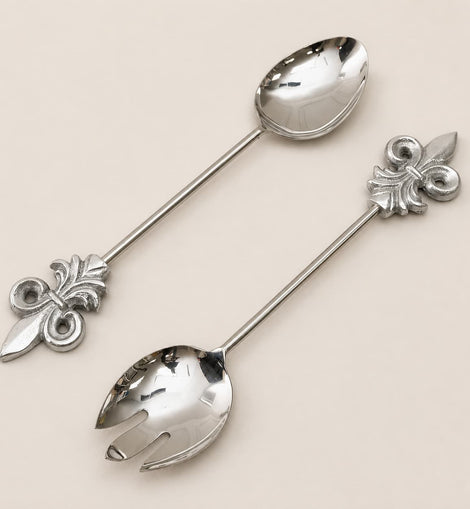 Two polished silver serving utensils with decorative fleur-de-lis handles, one spoon and one fork, arranged diagonally on a light beige background.