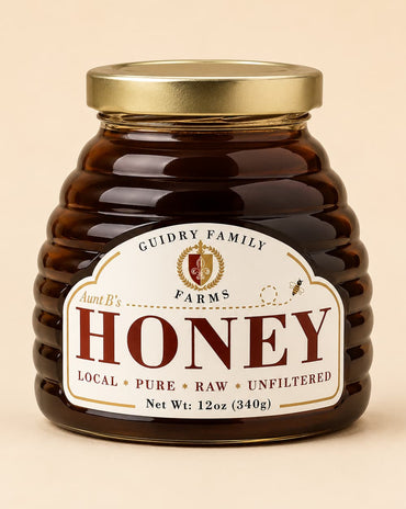 A jar of honey from Guidry Family Farms, Aunt B's, with a ribbed amber glass body, a gold metal lid, and a cream-colored label with red lettering, set against a solid beige background.