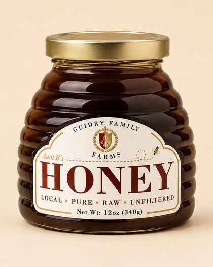A jar of honey from Guidry Family Farms, Aunt B's, with a ribbed amber glass body, a gold metal lid, and a cream-colored label with red lettering, set against a solid beige background.