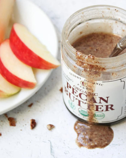 A jar of Organic Pecan Butter with a spoon inside, shown next to a white plate of sliced red apples on a light textured surface.
