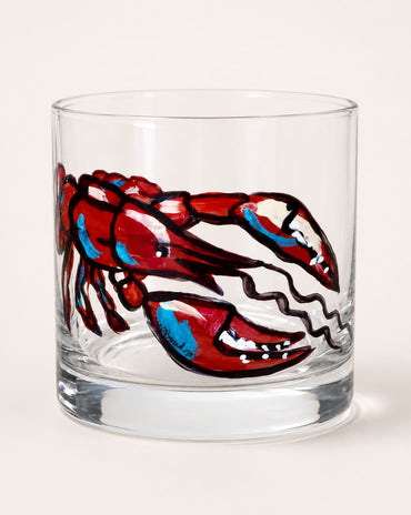 A clear glass rocks tumbler featuring a hand-painted lobster design in red, blue, and white with black outlines, set against a plain light beige background.