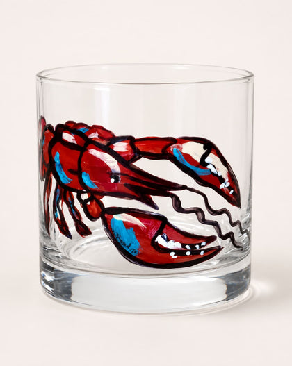 A clear glass rocks tumbler featuring a hand-painted lobster design in red, blue, and white with black outlines, set against a plain light beige background.