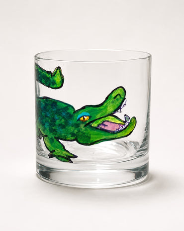A clear glass rocks tumbler featuring a hand-painted green alligator illustration on the side, set against a plain white background.