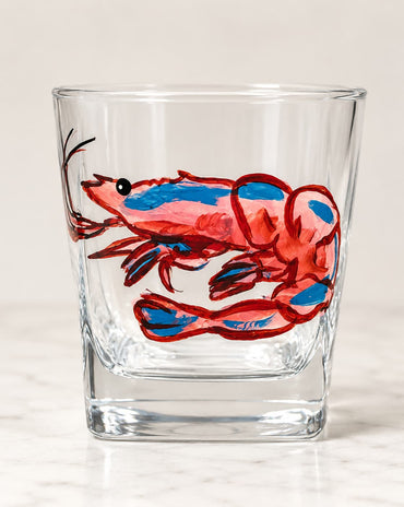 A clear glass tumbler with a hand-painted red and blue shrimp on the side, set against a light beige background.