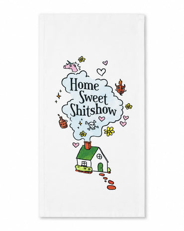 A white kitchen towel featuring a whimsical cartoon illustration of a small green house with a red chimney, emitting a light blue cloud of text that reads 'Home Sweet Shitshow' in black script. The design is decorated with small pink hearts, yellow flowers, and other doodles, set against a clean white background.