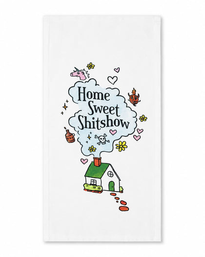 A white kitchen towel featuring a whimsical cartoon illustration of a small green house with a red chimney, emitting a light blue cloud of text that reads 'Home Sweet Shitshow' in black script. The design is decorated with small pink hearts, yellow flowers, and other doodles, set against a clean white background.