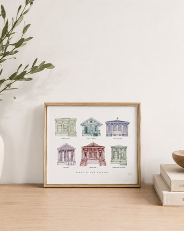 A framed art print featuring six vintage architectural sketches of New Orleans houses, styled on a light wood table next to a white vase with greenery and a stack of books.