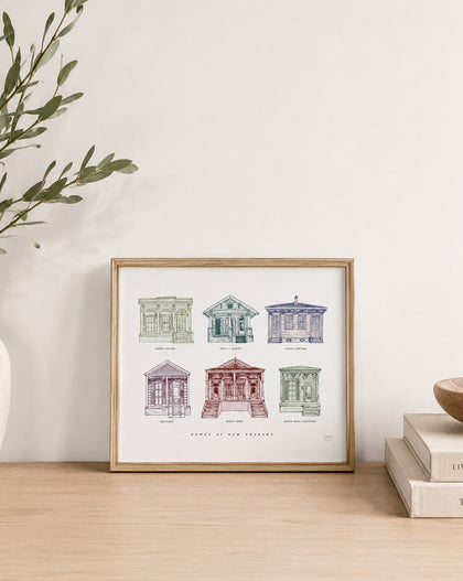 A framed art print featuring six vintage architectural sketches of New Orleans houses, styled on a light wood table next to a white vase with greenery and a stack of books.