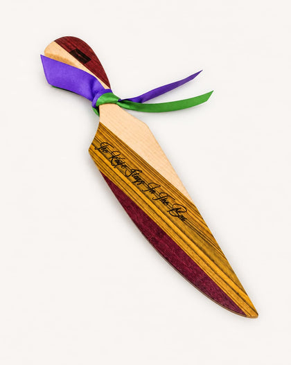A wooden king cake knife with a striped wood-grain finish, featuring black cursive engraving on the blade and tied with purple, green satin ribbons, shown on a plain off-white background.