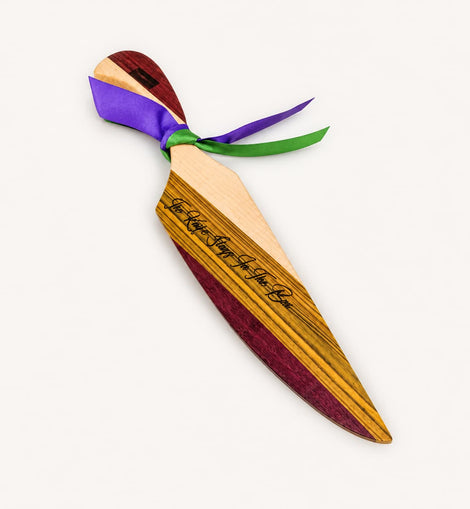 A wooden king cake knife with a striped wood-grain finish, featuring black cursive engraving on the blade and tied with purple, green satin ribbons, shown on a plain off-white background.