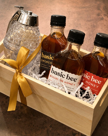 A wooden gift box containing three bottles of Basic Bee honey and a clear glass honey dispenser tied with a gold ribbon, set on a brown countertop.
