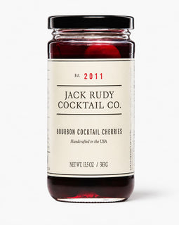 A glass jar of Jack Rudy Cocktail Co. Bourbon Cocktail Cherries with a cream-colored label and a black lid, set against a plain light gray background.