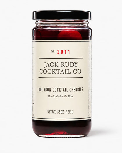 A glass jar of Jack Rudy Cocktail Co. Bourbon Cocktail Cherries with a cream-colored label and a black lid, set against a plain light gray background.