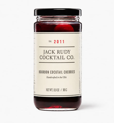A glass jar of Jack Rudy Cocktail Co. Bourbon Cocktail Cherries with a cream-colored label and a black lid, set against a plain light gray background.