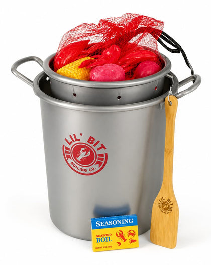 A  fake stainless steel pot set with a red mesh bag of red and yellow items on top, a wooden spoon, and a seasoning packet on a white background. The pot has a red circular logo that reads 'Lil' Bit Boiling Co.
