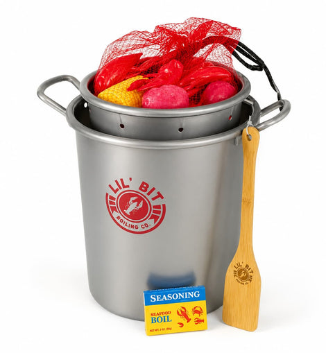 A  fake stainless steel pot set with a red mesh bag of red and yellow items on top, a wooden spoon, and a seasoning packet on a white background. The pot has a red circular logo that reads 'Lil' Bit Boiling Co.