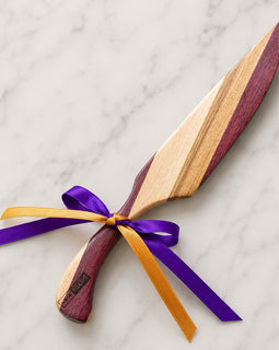 A wooden serving knife with a dual-tone wood blade and handle, tied with a purple and gold ribbon, displayed on a white marble background.