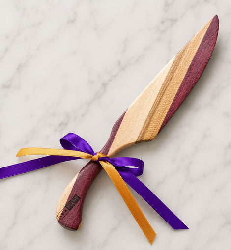 A wooden serving knife with a dual-tone wood blade and handle, tied with a purple and gold ribbon, displayed on a white marble background.