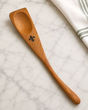 A wooden roux spoon with a black fleur-de-lis engraving, placed diagonally on a white marble surface next to a white kitchen towel with green stripes.