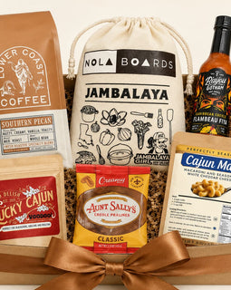 Gift basket  filled with multiple branded food items: a brown coffee bag, a cream drawstring bag labeled Jambalaya, a bottle of Camboise Pit sauce, a bag of Cajun Mac seasoning, and a pouch of Aunt Sally's Creole Prawns, arranged in a wooden tray with shredded filler and a large brown ribbon bow.
