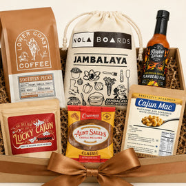 Gift basket  filled with multiple branded food items: a brown coffee bag, a cream drawstring bag labeled Jambalaya, a bottle of Camboise Pit sauce, a bag of Cajun Mac seasoning, and a pouch of Aunt Sally's Creole Prawns, arranged in a wooden tray with shredded filler and a large brown ribbon bow.