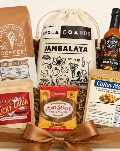 Gift basket  filled with multiple branded food items: a brown coffee bag, a cream drawstring bag labeled Jambalaya, a bottle of Camboise Pit sauce, a bag of Cajun Mac seasoning, and a pouch of Aunt Sally's Creole Prawns, arranged in a wooden tray with shredded filler and a large brown ribbon bow.