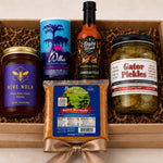 A curated gift box containing five distinct food products arranged in brown crinkle paper inside a light wood box. The items include a jar of Hive Nola honey, a blue and purple canister of Willes Fruit of the Loom, a bottle of Lanreau Fields habanero pepper sauce, a bag of Cajun Marinated shrimp, and a jar of Gator Pickles.