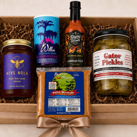 A curated gift box containing five distinct food products arranged in brown crinkle paper inside a light wood box. The items include a jar of Hive Nola honey, a blue and purple canister of Willes Fruit of the Loom, a bottle of Lanreau Fields habanero pepper sauce, a bag of Cajun Marinated shrimp, and a jar of Gator Pickles.