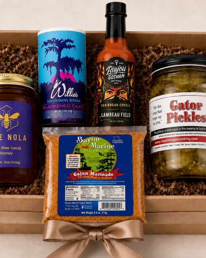 A curated gift box containing five distinct food products arranged in brown crinkle paper inside a light wood box. The items include a jar of Hive Nola honey, a blue and purple canister of Willes Fruit of the Loom, a bottle of Lanreau Fields habanero pepper sauce, a bag of Cajun Marinated shrimp, and a jar of Gator Pickles.