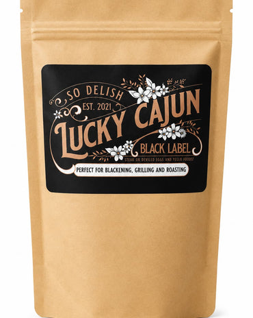 A kraft paper stand-up pouch with a black label reading 'So Delish Lucky Cajun Black Label' and decorative white floral details, shown on a white background.