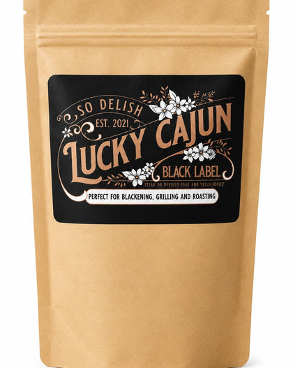 A kraft paper stand-up pouch with a black label reading 'So Delish Lucky Cajun Black Label' and decorative white floral details, shown on a white background.