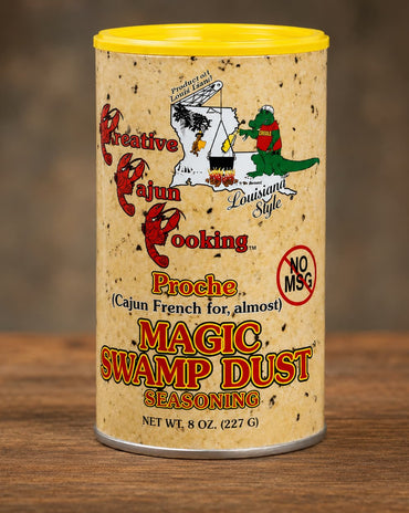 A 8 oz canister of Magic Swamp Dust Seasoning on a wooden surface with a blurred brown background. The cylindrical container has a yellow lid, a beige speckled label with Cajun-themed illustrations, and red and orange text.