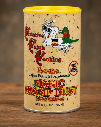 A 8 oz canister of Magic Swamp Dust Seasoning on a wooden surface with a blurred brown background. The cylindrical container has a yellow lid, a beige speckled label with Cajun-themed illustrations, and red and orange text.