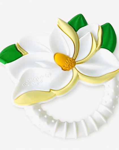 A white silicone baby teether with a magnolia flower design, featuring gold-colored accents and green leaves, on a light off-white background.