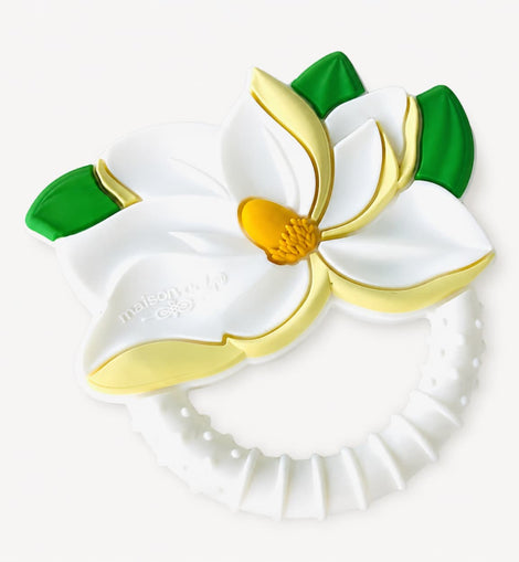 A white silicone baby teether with a magnolia flower design, featuring gold-colored accents and green leaves, on a light off-white background.