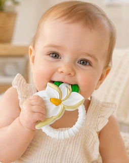 A baby wearing a cream ribbed sleeveless top holds a white magnolia shaped teether with green and yellow accents in their mouth.