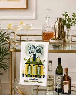 A white kitchen towel with a colorful New Orleans-themed illustration and the text 'Make mine a Double!' is draped over a gold bar cart. The scene is styled with cocktail glasses, a decanter, a shaker, and liquor bottles on a glass bar table against a white paneled wall.