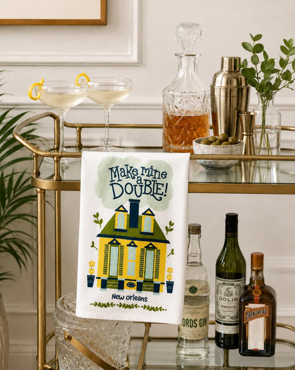 A white kitchen towel with a colorful New Orleans-themed illustration and the text 'Make mine a Double!' is draped over a gold bar cart. The scene is styled with cocktail glasses, a decanter, a shaker, and liquor bottles on a glass bar table against a white paneled wall.