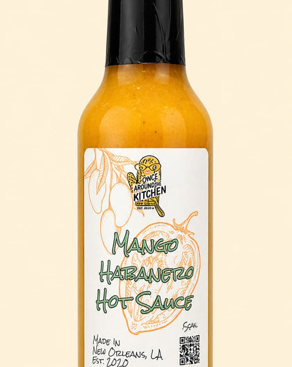 A bottle of Mango Habanero Hot Sauce by The Sweet Kitchen, featuring a black cap and a white label with green and yellow text, set against a solid pale beige background.