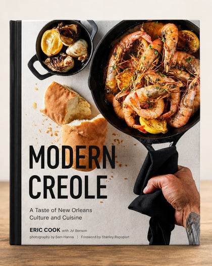 The front cover of the book 'Modern Creole' by Eric Cook with Jil Benson is shown standing upright on a wooden surface. The cover features a large photograph of shrimp and seafood in a cast-iron skillet, bread, and a small pot, with the title in bold black text on a light background.