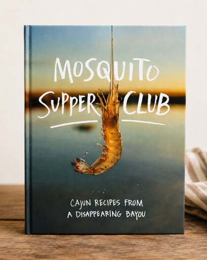 A hardcover book titled 'Mosquito Supper Club' with a cover photo of a shrimp hanging from a hook over water, standing on a wooden surface.