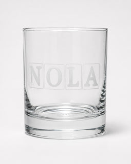A clear glass rocks tumbler with the name 'NOLA' printed in a frosted block-style font on the front, set against a plain light gray background.