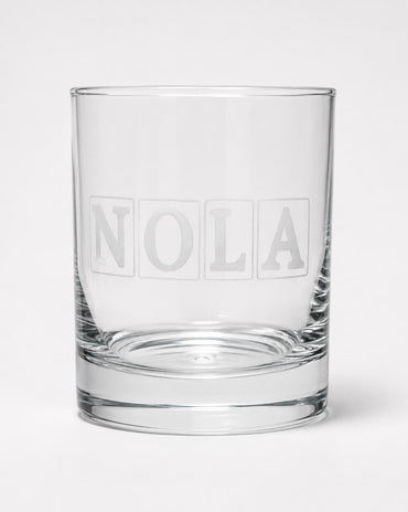 A clear glass rocks tumbler with the name 'NOLA' printed in a frosted block-style font on the front, set against a plain light gray background.