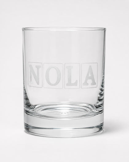 A clear glass rocks tumbler with the name 'NOLA' printed in a frosted block-style font on the front, set against a plain light gray background.